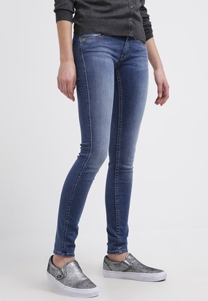 Osoba ubrana w niebieskie obcisłe dżinsy, ciemnoszary top z długim rękawem i srebrne wsuwane trampki, stojąca na prostym białym tle.