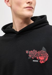 Νεαρό άτομο που φοράει μαύρη φούτερ με κουκούλα με κόκκινο γραφικό τριαντάφυλλο και κείμενο «HUGO» στο στήθος, κοιτάζοντας ελαφρώς προς τα δεξιά, ουδέτερο φόντο.