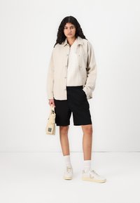 Beige linned skjortejakke, hvid kabelstrikket sweater, sorte shorts, beige sneakers med accenter, holder en lille lysebrun taske. Enkel baggrund.