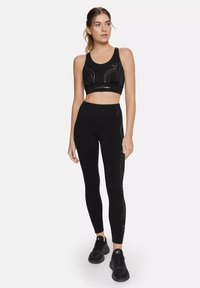 Crni sportski crop top s zlatnim linijama, uparen s crnim tajicama koje imaju slične zlatne akcentne detalje. Mekani, rastezljivi materijal, prilagodljiv kroj.