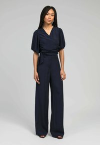 Femme avec de longues tresses portant un haut bleu marine à manches bouffantes et un pantalon large à taille haute, se tenant devant un fond uni.