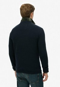 Marineblauer Strickpullover mit strukturierter Oberfläche und grünem Karokragen, kombiniert mit blauen Jeans. Ansicht von hinten.