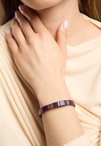 Pulsera morada con diseño rectangular, acabado brillante y reluciente, con detalles metálicos, descansando sobre una muñeca desnuda contra una tela suave y de tono neutro.