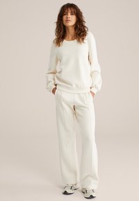 Mujer de pie con las manos en los bolsillos, vistiendo un sudadera de color crema, pantalones de pierna ancha a juego y zapatillas beige, sobre un fondo neutral.