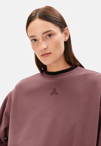 Junge Frau mit glattem braunem Haar und Sommersprossen, die einen mauvefarbenen Sweatshirt mit einem kleinen gestickten Logo auf einem hellen Hintergrund trägt.
