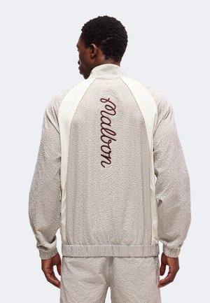 Homme portant une veste texturée beige et un pantalon assorti, vue de dos, avec "woolrich" brodé verticalement sur la veste.