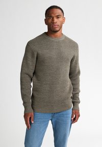 Olijfgroen gebreid sweater met een textuurpatroon, geribbelde manchetten en zoom. In combinatie met lichtblauwe jeans staat het model met een neutrale uitdrukking.