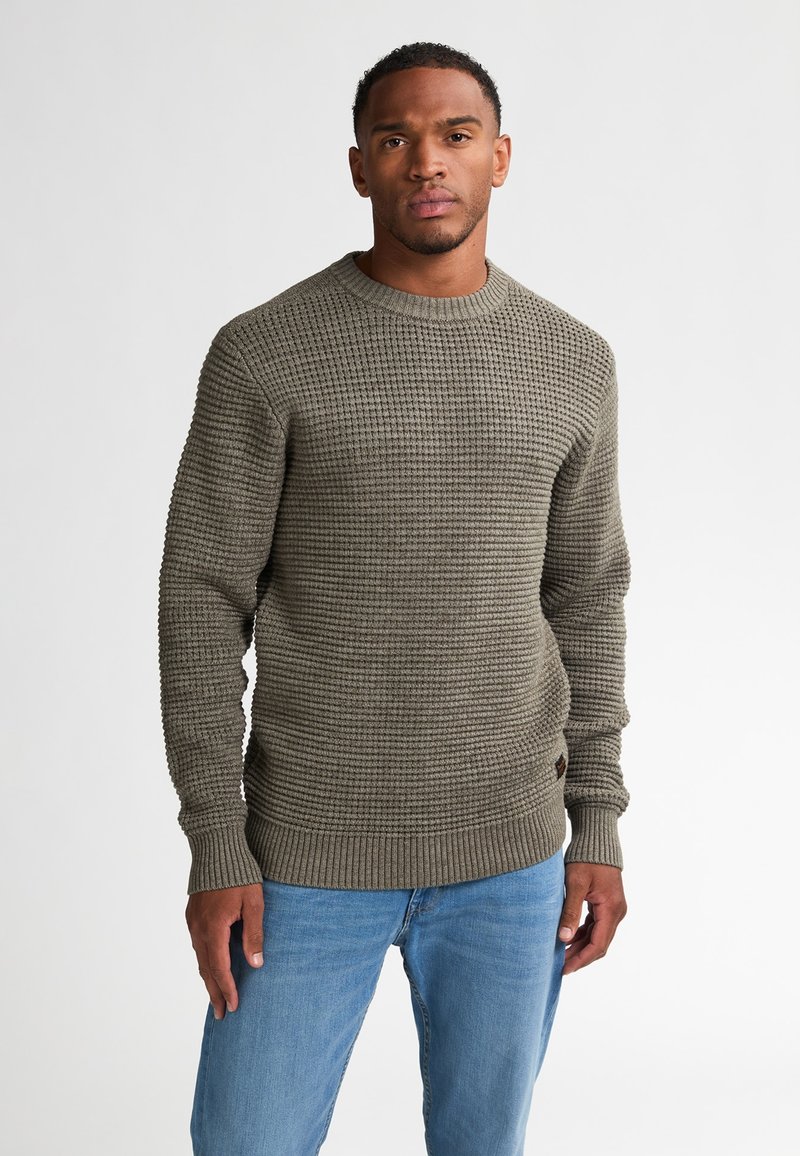Olijfgroen gebreid sweater met een textuurpatroon, geribbelde manchetten en zoom. In combinatie met lichtblauwe jeans staat het model met een neutrale uitdrukking.