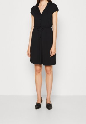 Vrouw draagt een zwarte korte mouws jurk tot op de knie met knopen en een taillekoord, gecombineerd met zwarte pumps met een lage hak en een spits toelopende neus.