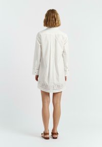 Abito camicia bianco in cotone, con maniche lunghe, colletto abbottonato e dettagli floreali ricamati all'orlo. Texture liscia.