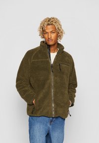 Giacca in pile verde oliva con chiusura a zip, colletto alto e tasche laterali. Presenta una superficie testurizzata e zip nere a contrasto.