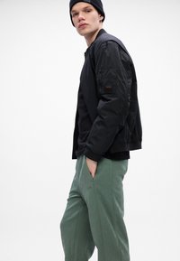 Schwarze Bomberjacke mit gerippten Bündchen über einem schwarzen Shirt. Olivgrüne, strukturierte Jogginghosen mit Logo, Hände in den Taschen, einfarbiger Hintergrund.