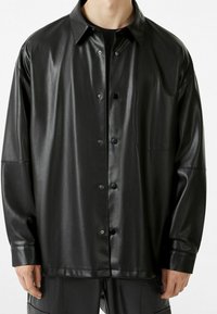 Camisa de cuero sintético negra con cierre de botones en el frente, corte holgado, cuello puntiagudo y un bolsillo en el pecho. Presenta una textura suave y un acabado brillante.