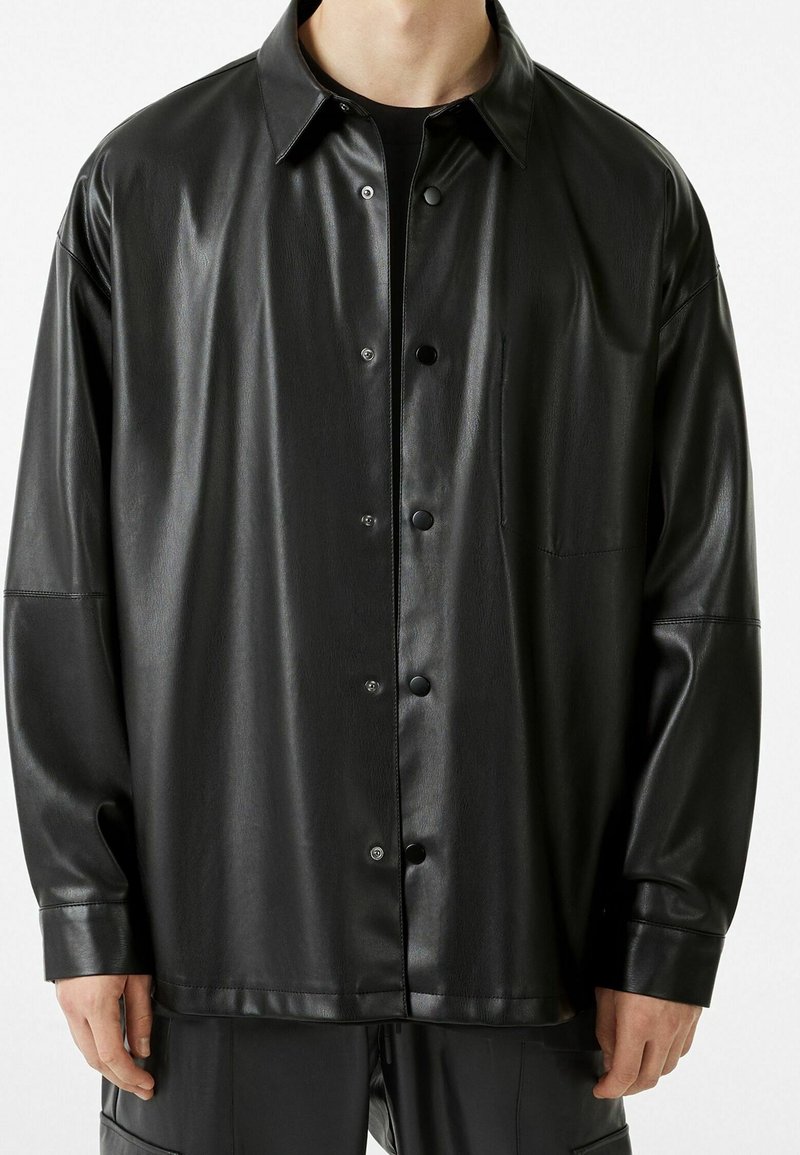 Camisa de cuero sintético negra con cierre de botones en el frente, corte holgado, cuello puntiagudo y un bolsillo en el pecho. Presenta una textura suave y un acabado brillante.