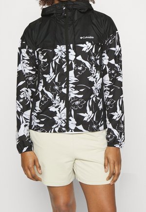 Sort-hvidt blomster Columbia-jakke med hætte, lynlås foran og elastiske manchetter, båret med lyse beige shorts.