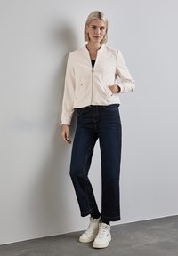 Giacca bomber rosa chiaro con zip frontale, tasche laterali e maniche a costine. Abbinata a jeans larghi blu scuro e sneakers bianche.