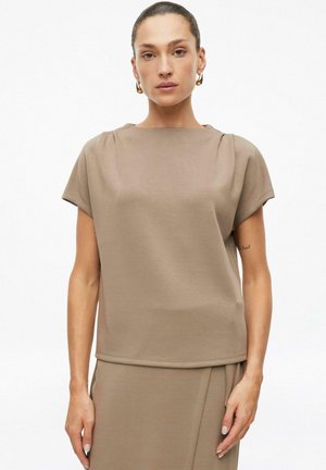 Vrouw met achter gebonden haar, die een beige top met korte mouwen en een bijpassende rok draagt tegen een effen witte achtergrond.