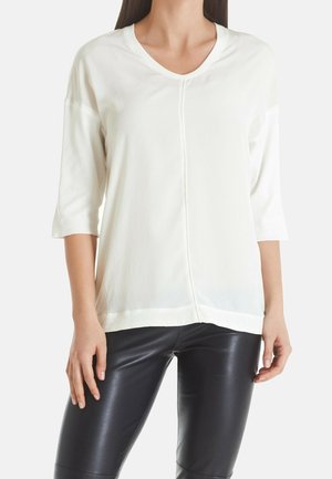 Femme portant un haut blanc à col en V avec des manches trois-quarts et des leggings en cuir noir, debout devant un fond uni.