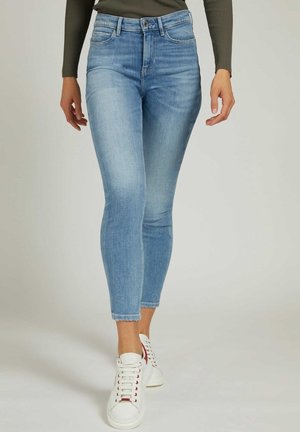 Kvinde iført lyseblå skinny jeans og hvide sneakers, vist fra taljen og ned mod fødderne mod en neutral baggrund.