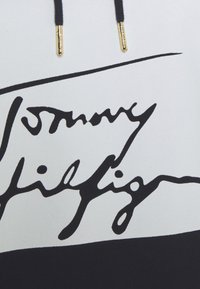 Cordons noirs avec embouts dorés au-dessus d'un texte noir en cursive sur tissu blanc, partie d'un sweat à capuche ou d'un sweat-shirt.