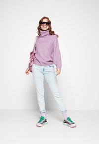 Frau lächelt, trägt übergroße Sonnenbrille, lilafarbene Rollkragenpullover, helle Jeans, bunte Sneakers und hält eine florale Tragetasche.