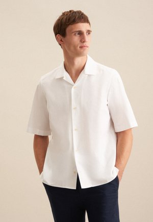 Chemise blanche à manches courtes en tissu léger, avec un col évasé, une fermeture à boutons et une texture subtile. Portée avec un pantalon bleu marine.