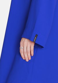 Helles blaues Kleidungsstück mit einer Detailierung am Ärmel. Eine Hand ruht auf dem Stoff und trägt einen silbernen herzförmigen Ring. Ein schwarzes Etikett ist in der Nähe des Ärmels sichtbar.