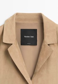Col de blazer en daim beige avec une étiquette noire portant l'inscription "Massimo Dutti" et un texte plus petit "Fabriqué en Inde" à l'intérieur du vêtement.