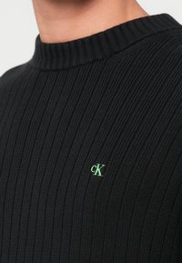 Čierny prúžkovaný sveter z pleteného materiálu, s vysokým golierom a malým zeleným logom na hrudi.