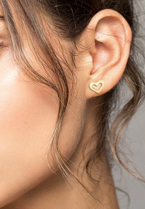 Goldene, herzförmige Stecker Ohrringe mit strukturierten Kanten, vor einer Nahaufnahme des Ohres eines Models und welligem Haar, das das zarte Design betont.