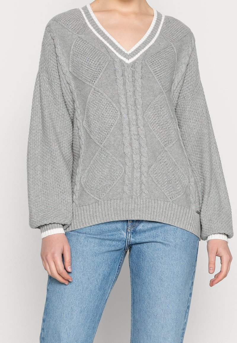 Femme portant un pull gris à col en V avec un motif diamant texturé, associé à un jean bleu clair, sur un fond uni.