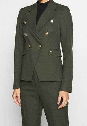 Mørkegrøn dobbeltradet blazer med guldknapper båret over matchende bukser og en sort top på en stående person.