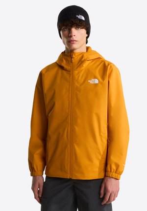 Oranje waterdichte jas met capuchon, elastische manchetten en een rits aan de voorkant. Heeft een logo op de borst. Model draagt een zwarte gebreide muts.