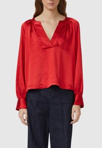 Frau trägt eine locker sitzende, glänzende rote Bluse mit langen Ärmeln und Schlitz am Ausschnitt sowie dunkle, hoch taillierte Hosen vor einem einfarbigen Hintergrund.