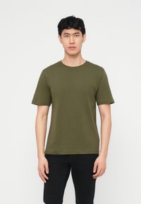 Young man with short black hair wearing a plain olive green short-sleeve T-shirt and black pants, standing against a white background.