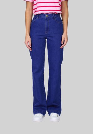 Personne portant un jean évasé taille haute bleu foncé, des baskets blanches et un t-shirt à manches courtes rayé rose et blanc sur fond gris.