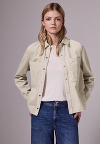 Veste chemise en velours côtelé beige clair avec deux poches poitrine et boutons-pression, associée à une chemise claire et un jean en denim bleu.