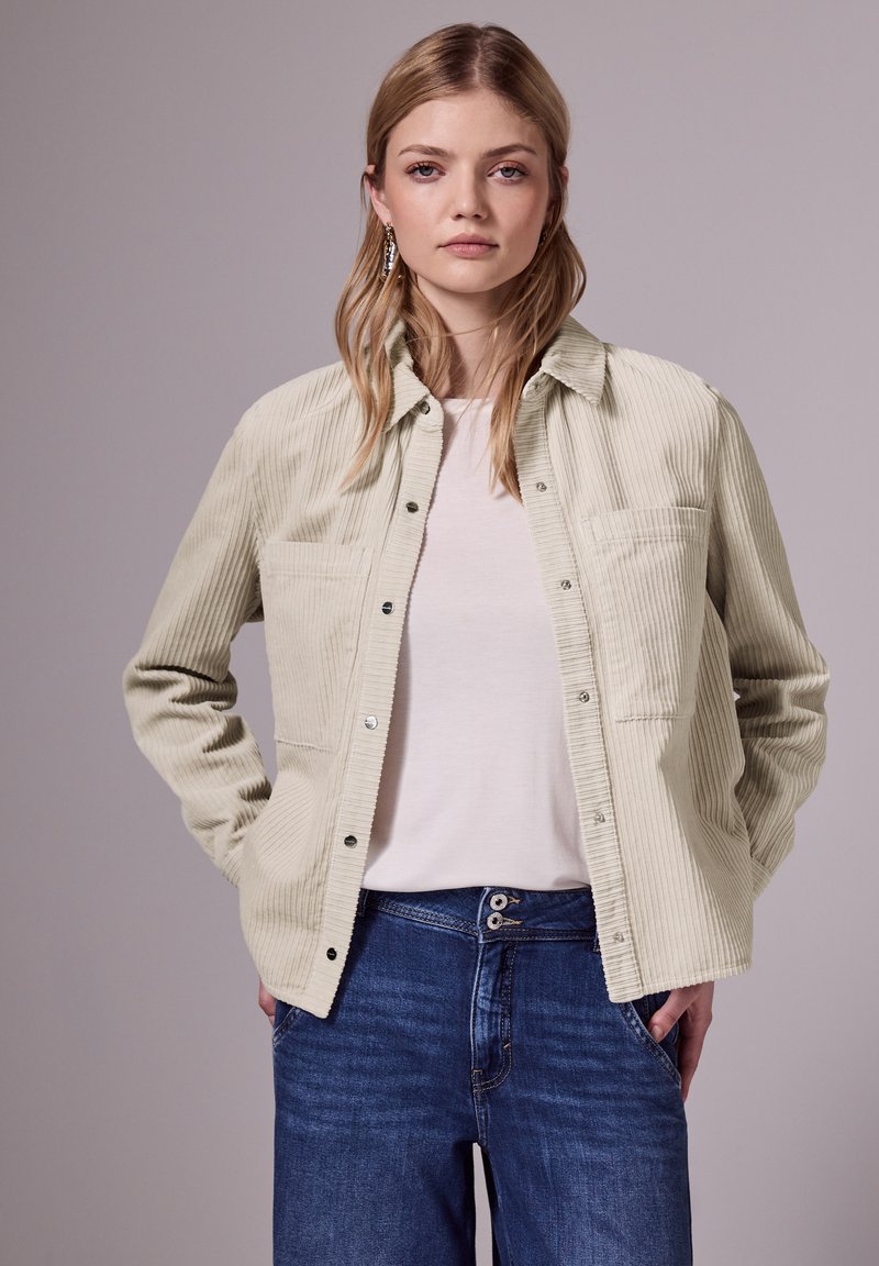 Veste chemise en velours côtelé beige clair avec deux poches poitrine et boutons-pression, associée à une chemise claire et un jean en denim bleu.
