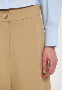 Beige weite Hosen mit glatter Textur, ausgestattet mit einem Knopfverschluss und vorderen Falten, kombiniert mit einem blauen Langarmhemd.