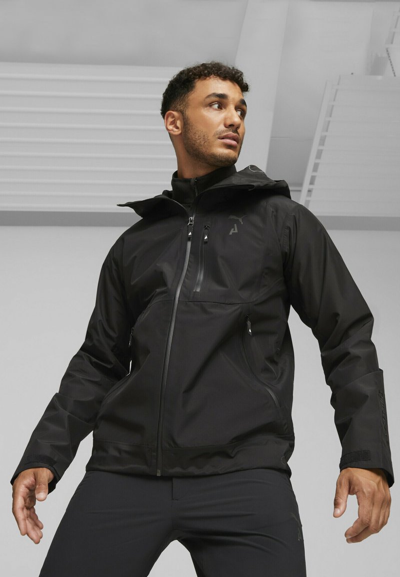Mann trägt eine schwarze Kapuzenjacke und schwarze Hose, steht drinnen mit leicht ausgestreckten Armen und schaut zur Seite.