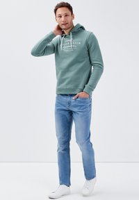 Grüne Kapuzenjacke mit weißem Text, Kapuze mit Zugband, gerippte Bündchen, kombiniert mit hellblauen Jeans und weißen Sneakers. Minimalistisches Design.
