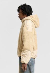 Beige fleecejacka med huva, strukturerad kropp och släta, ljusare paneldetaljer på ärmar och axlar.