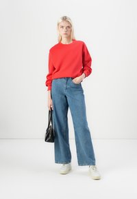 Červená mikina s dlhými rukávmi, kombinovaná so širokými modrými džínsami. Biely tenisky a malá čierna kabelka uzatvárajú outfit.