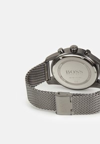 Montre en acier inoxydable noir avec un bracelet en maille, fonctions chronographe et boîtier rond. Logo gravé sur le fermoir du bracelet et au dos du boîtier.
