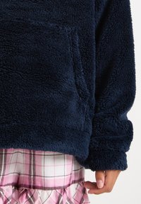 Navy blue, fuzzy pullover with a front pocket and ribbed cuffs, paired with pink and white plaid pajama shorts featuring a ruffled hem.