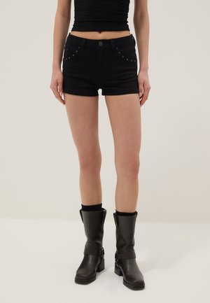 Frau trägt schwarze, hoch taillierte Jeansshorts mit Metallnieten, ein schwarzes Croptop und schwarze Lederstiefel bis zur Mitte der Wade und steht auf weißem Boden.