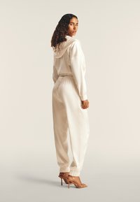 Hoodie in satin crema con polsini elasticizzati, abbinato a pantaloni tapered coordinati. Presenta un cappuccio con cordoncino e una vita slim. Sandali con tacco beige.