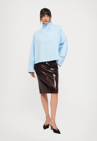 Woman standing wearing a light blue high-neck top with wide sleeves and a dark brown glossy pencil skirt, paired with black heels.