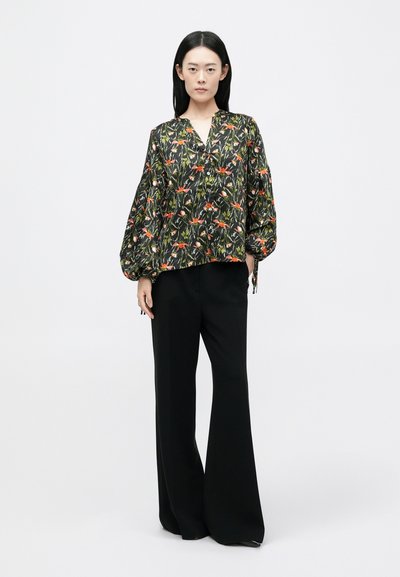 Juodas gėlių bluzonas su oranžiniais ir žaliais raštais, turintis V formos iškirptę ir pūstus rankovės, derinamas su plačiomis juodomis kelnėmis.