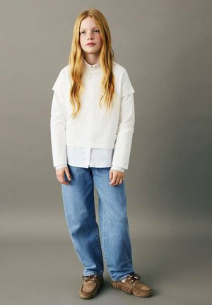 Haut blanc à manches longues superposé à une chemise à col, associé à un jean bleu clair à jambes larges et des mocassins marron.